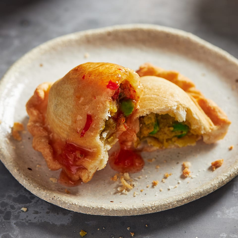 A golden-brown British Embassy pasty, broken open to reveal a savory filling, drizzled with sweet chili sauce.
