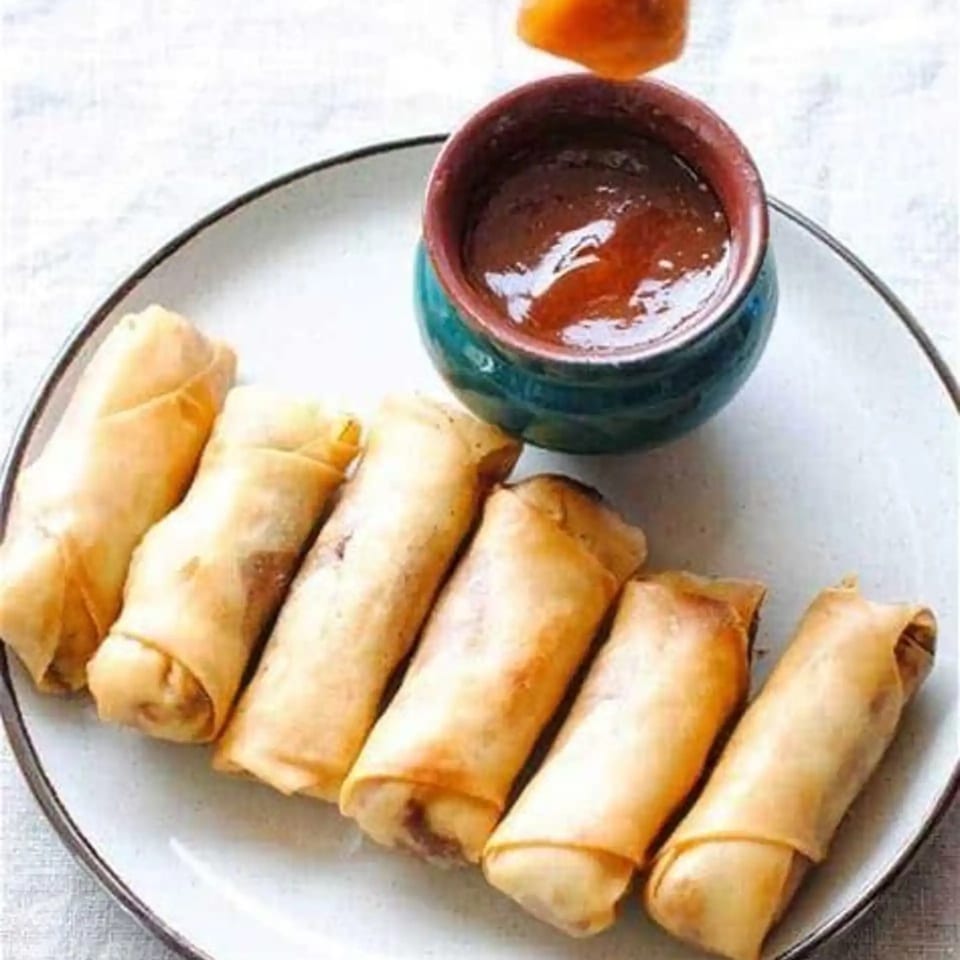Six golden spring rolls arranged on a white plate with a small teal bowl of dipping sauce.
