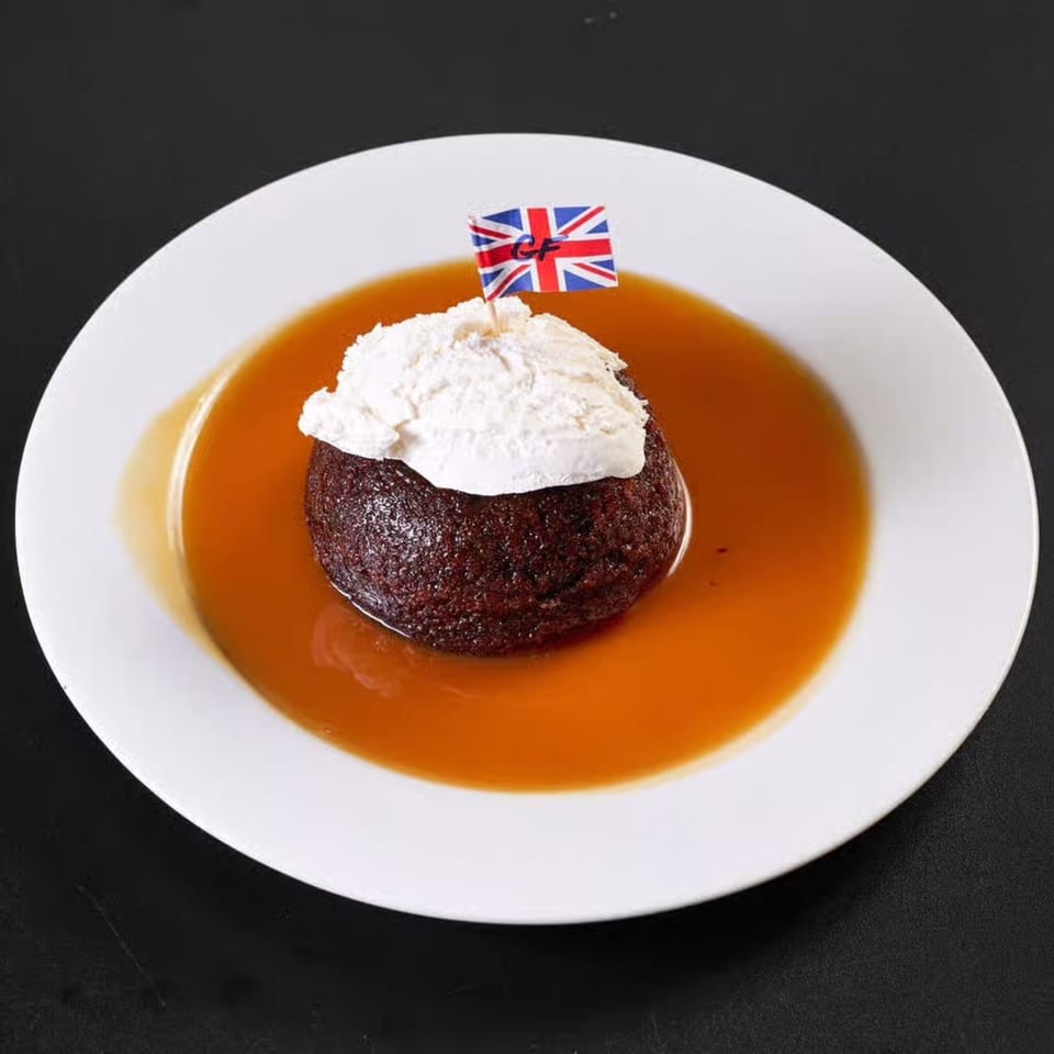 Sticky toffee pudding with whipped cream and a British flag on a white plate with caramel sauce.