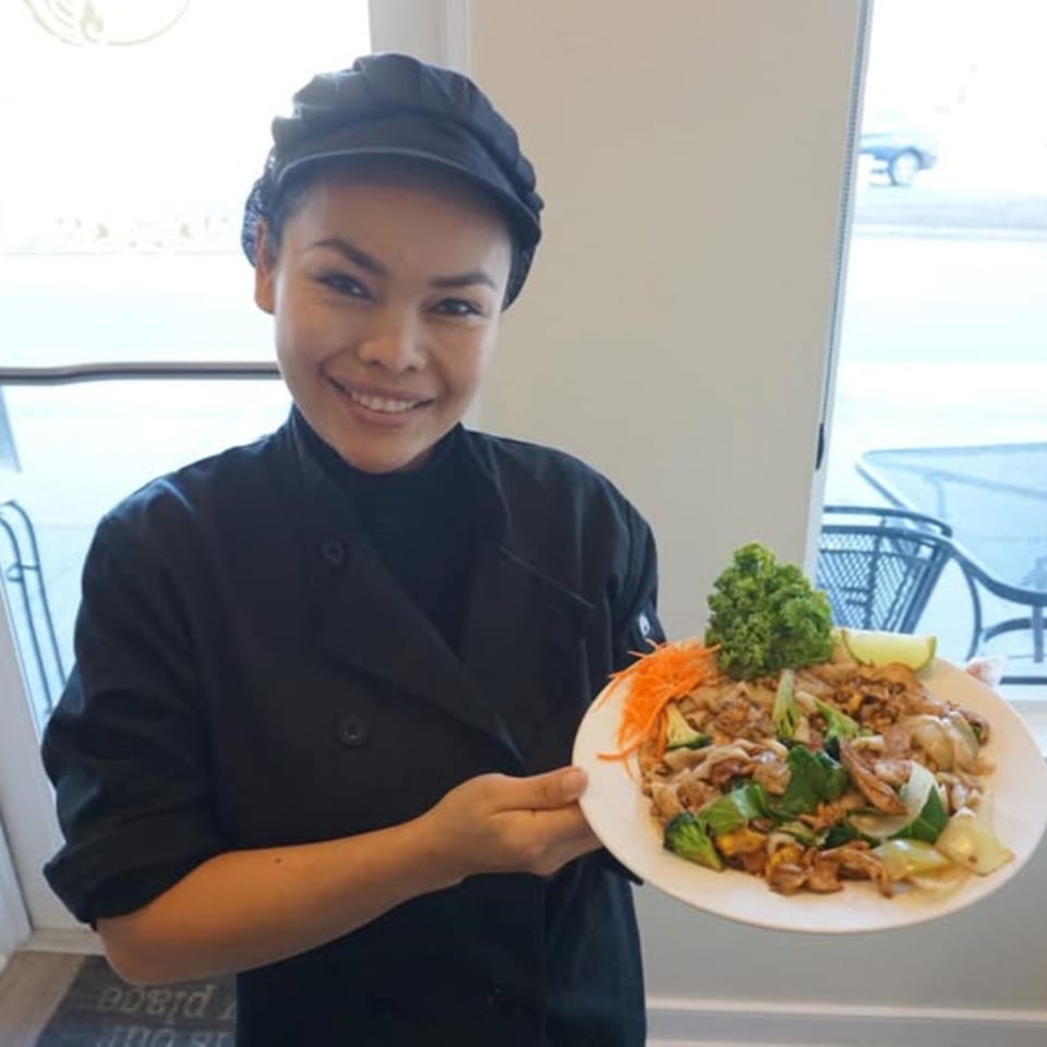 Smiling chef in black uniform holding a plate of stir-fried noodles with vegetables and meat.