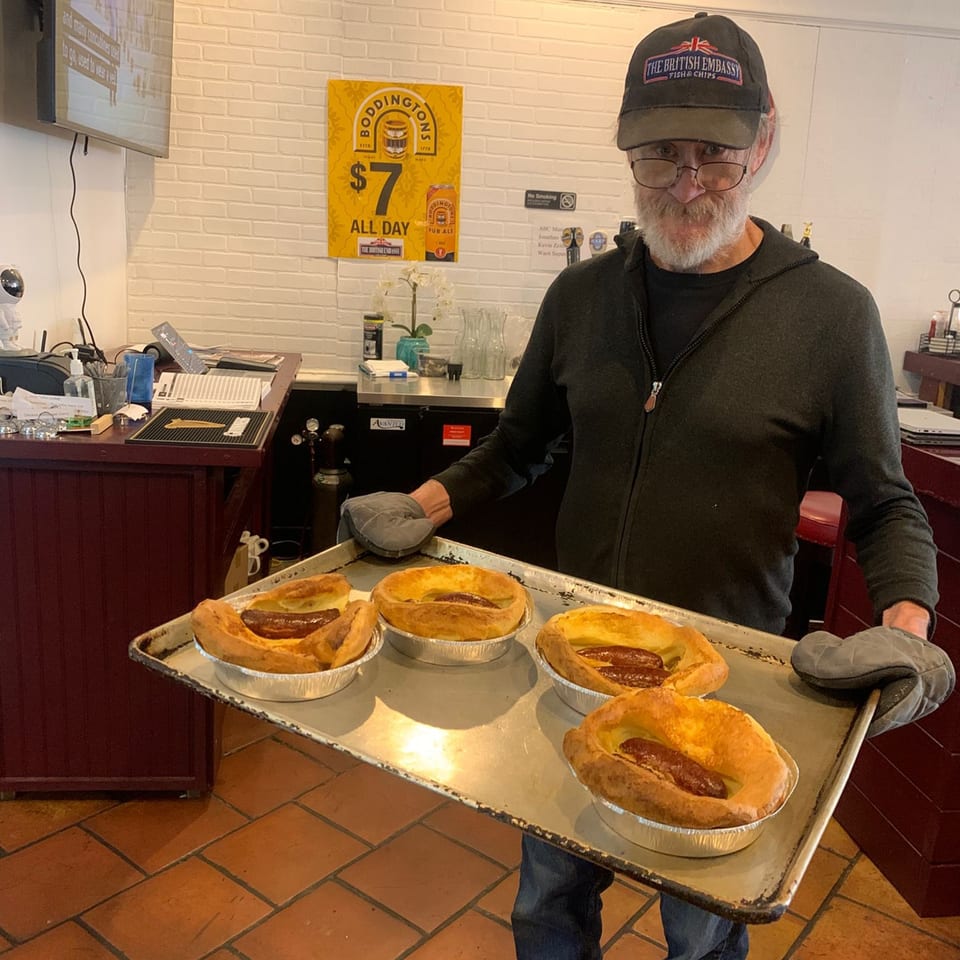 A man in a British Embassy hat holds a tray of four Toad in the Hole dishes.