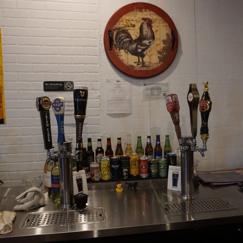 A bar counter with beer taps, bottled beers, and canned drinks, under a rooster wall decoration.
