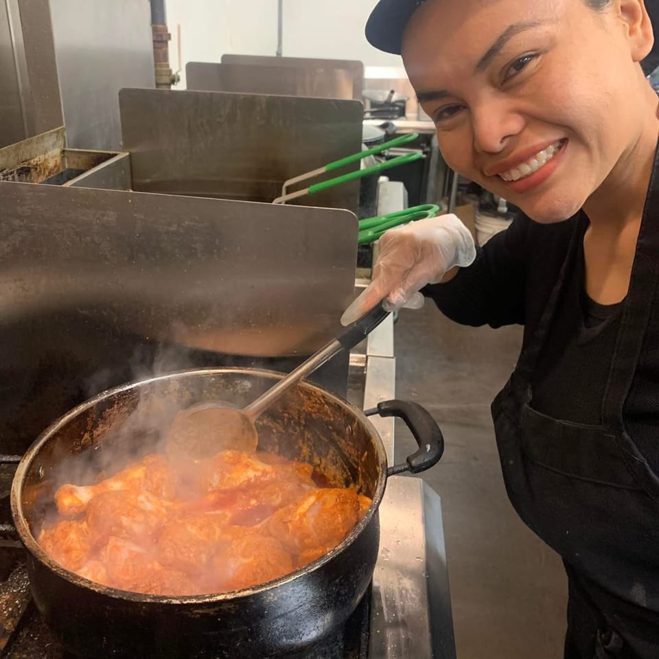 Smiling chef stirring a large pot of simmering chicken in a commercial kitchen.