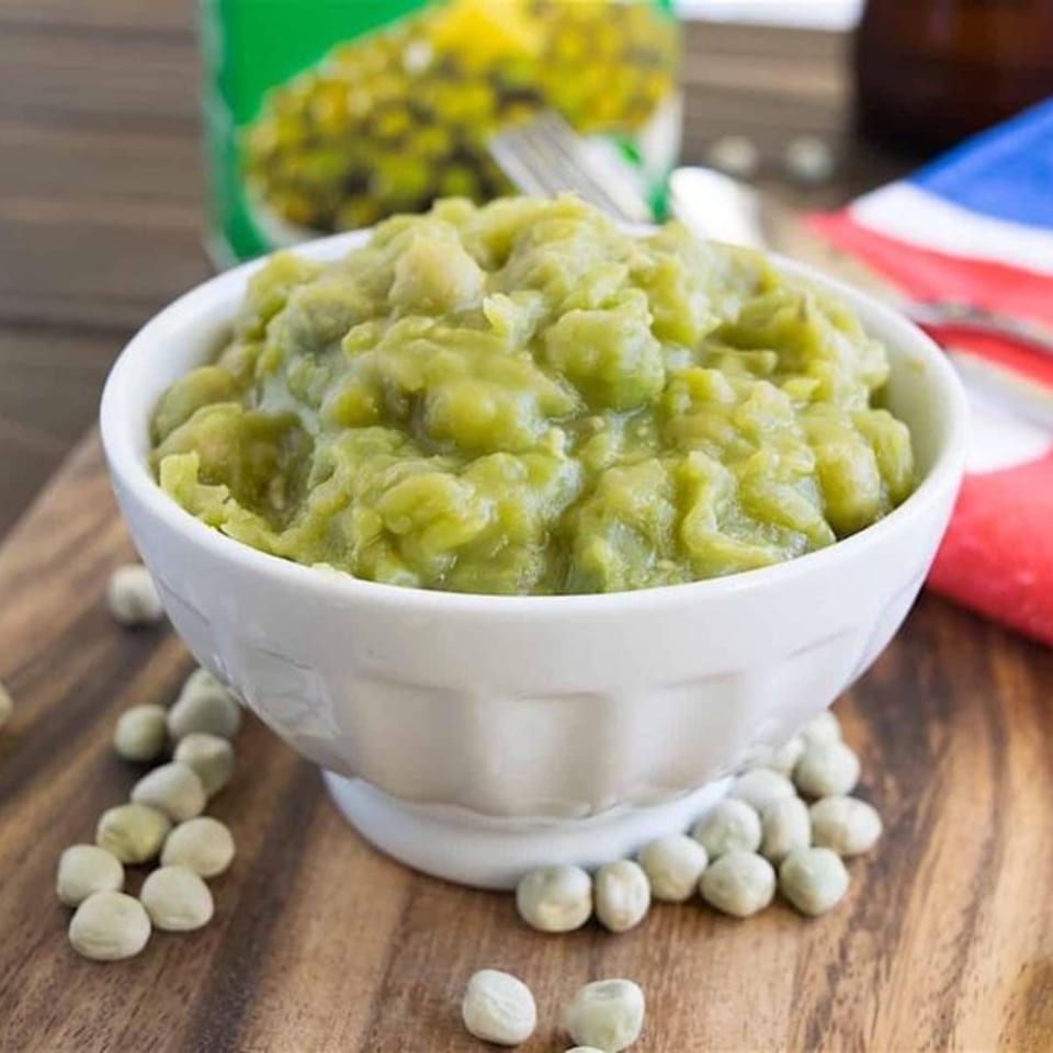 A white bowl of bright green mushy peas sits on a wooden board with scattered dried peas.